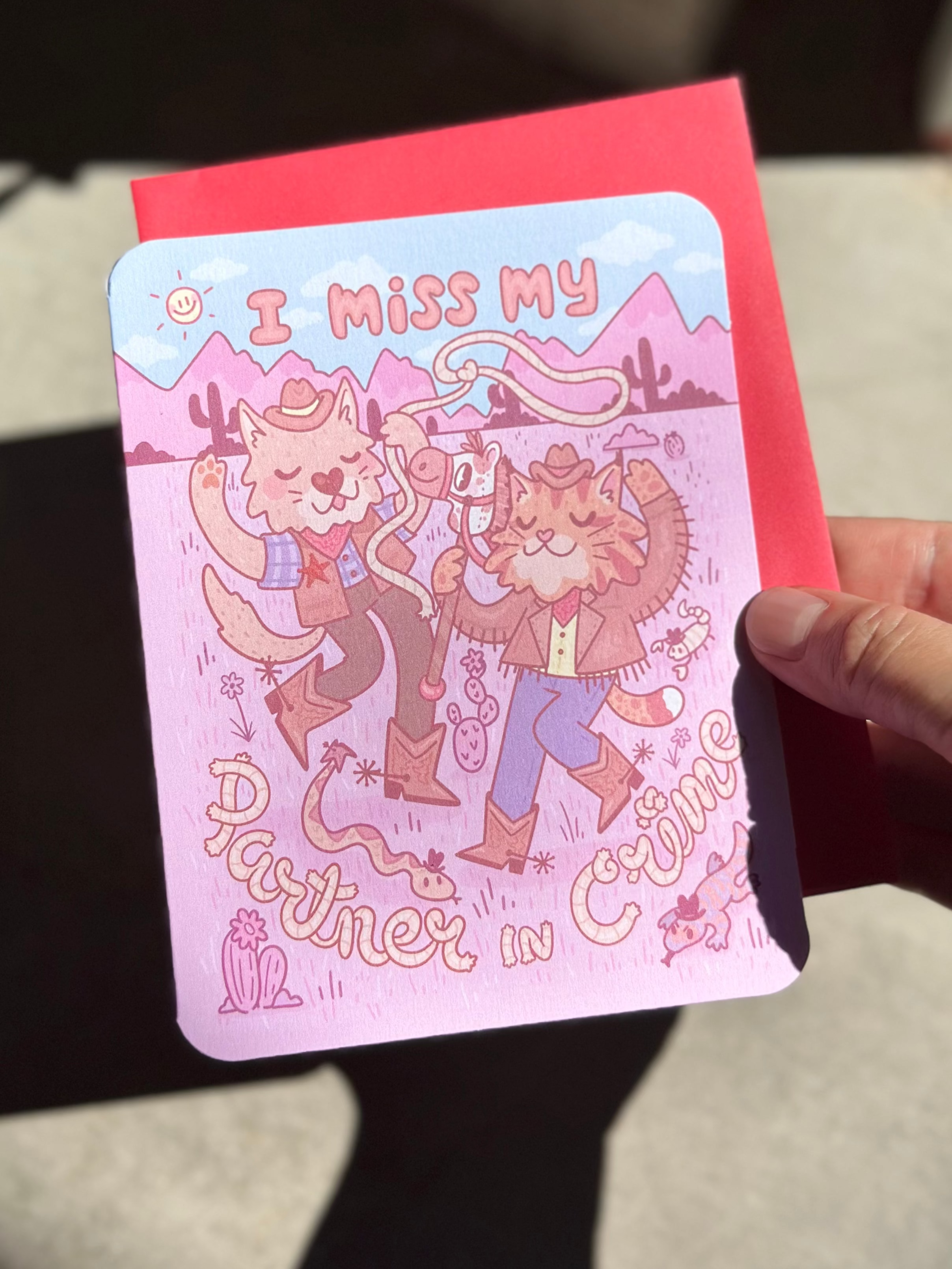 Pink western-themed greeting card with two cartoon cowboy cats dancing in the desert, wearing boots and hats, surrounded by cacti and snakes, with text ‘I miss my partner in crime,’ paired with red envelope.