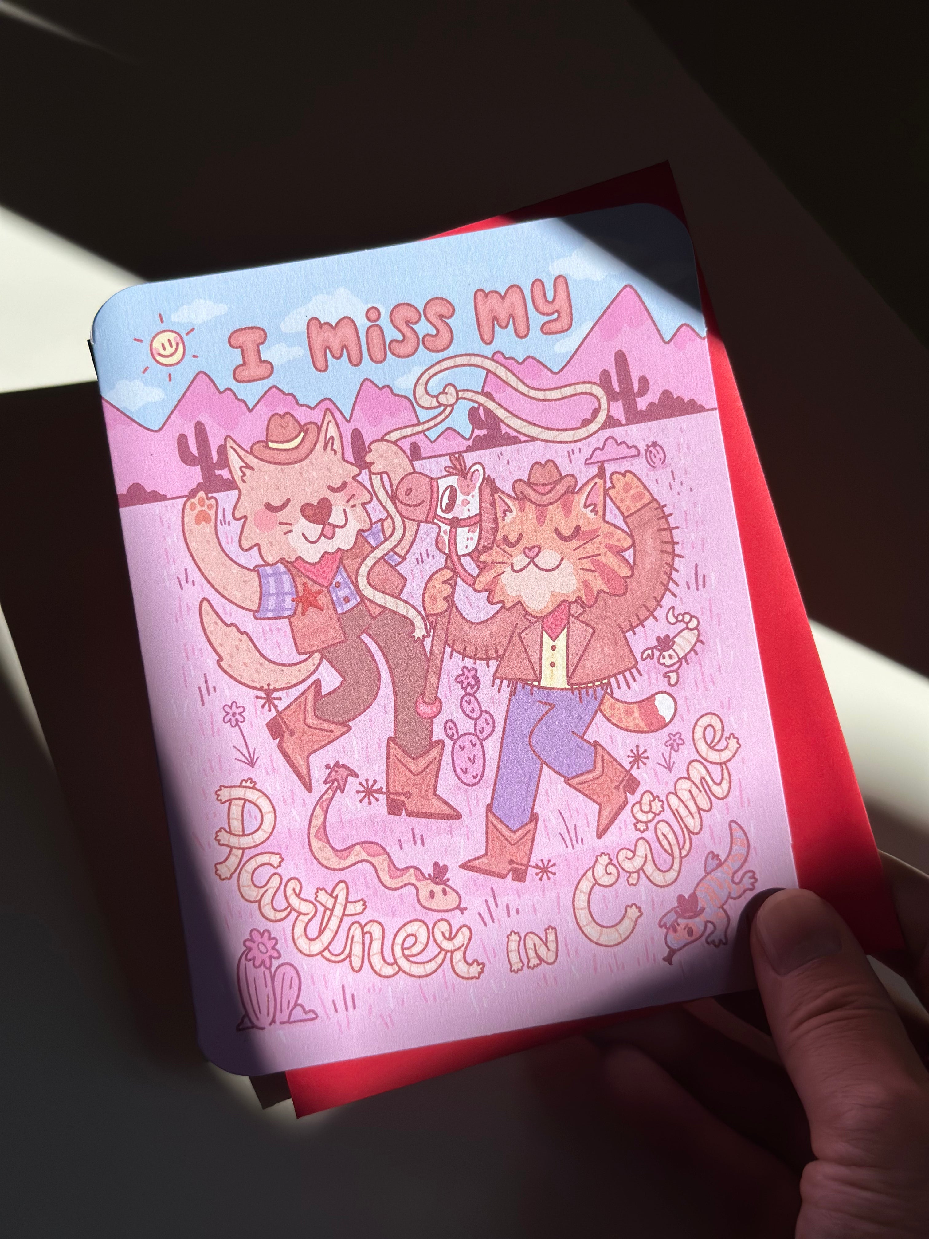 Pink western-themed greeting card with two cartoon cowboy cats dancing in the desert, wearing boots and hats, surrounded by cacti and snakes, with text ‘I miss my partner in crime,’ paired with red envelope.
