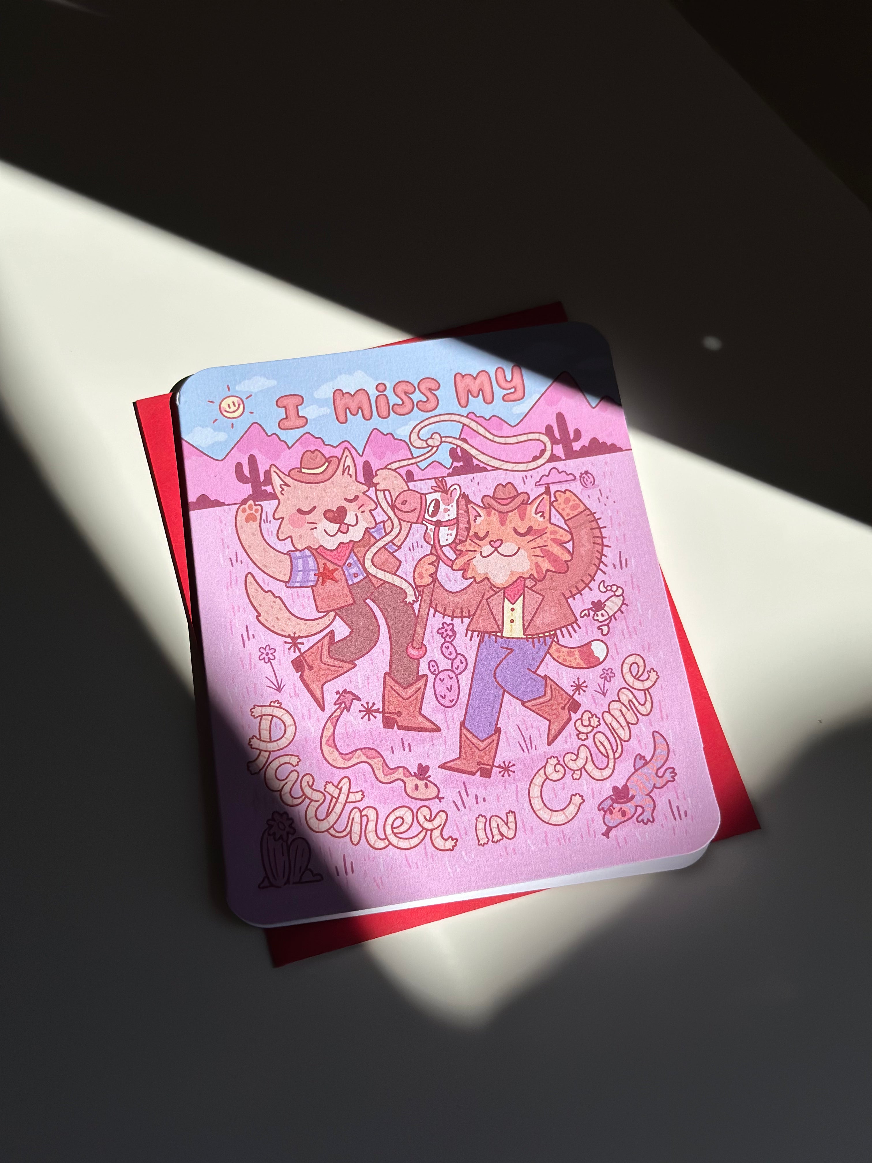 Pink western-themed greeting card with two cartoon cowboy cats dancing in the desert, wearing boots and hats, surrounded by cacti and snakes, with text ‘I miss my partner in crime,’ paired with red envelope.