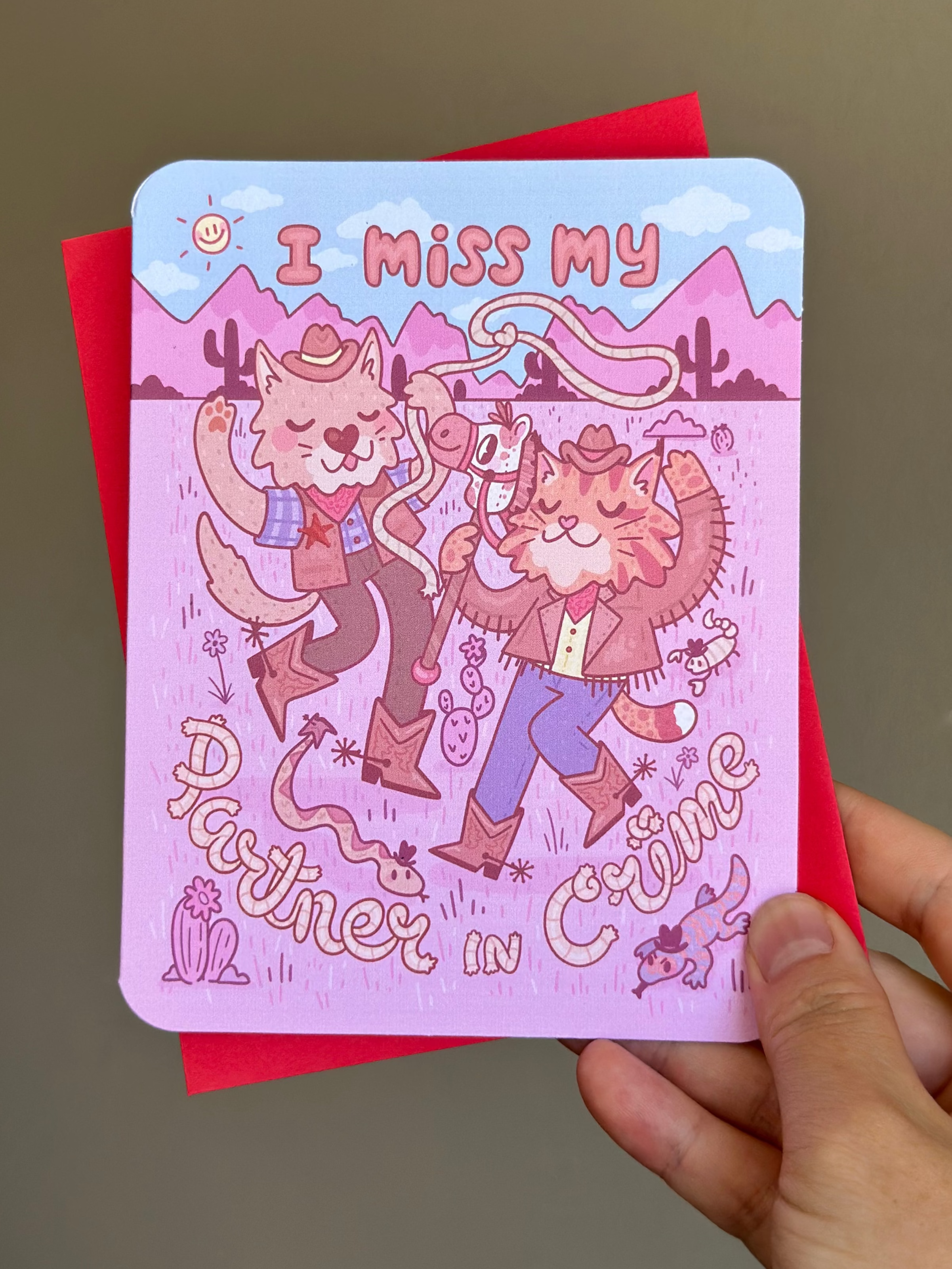 Pink western-themed greeting card with two cartoon cowboy cats dancing in the desert, wearing boots and hats, surrounded by cacti and snakes, with text ‘I miss my partner in crime,’ paired with red envelope.