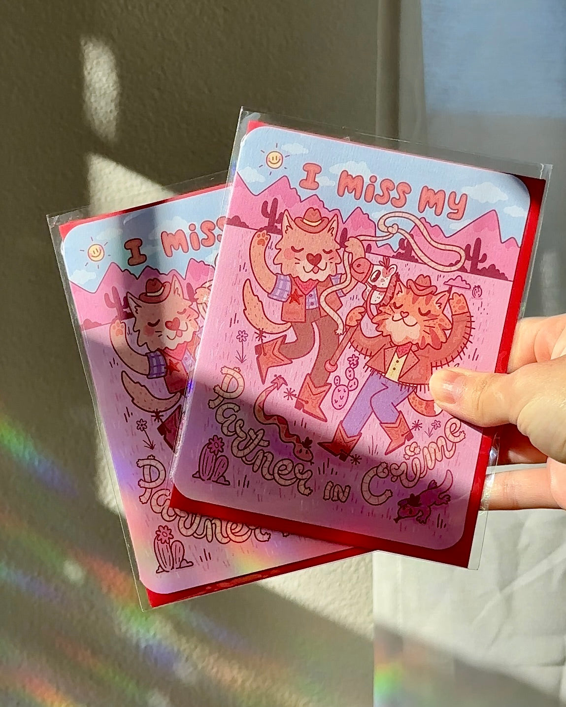 Pink western-themed greeting card with two cartoon cowboy cats dancing in the desert, wearing boots and hats, surrounded by cacti and snakes, with text ‘I miss my partner in crime,’ paired with red envelope.