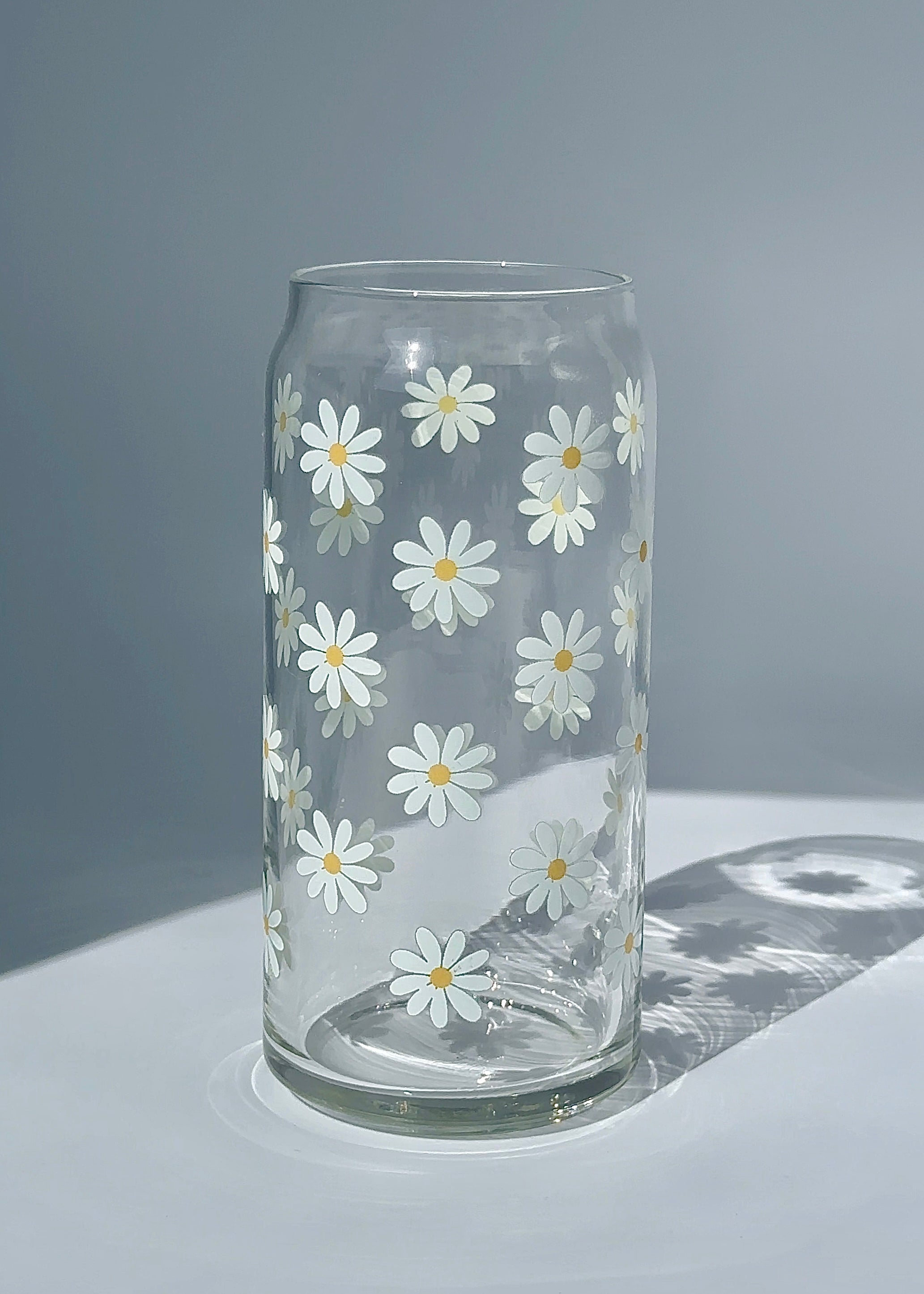 Clear 20 oz glass can with a minimalist white daisy pattern, styled in soft natural light on a clean surface.