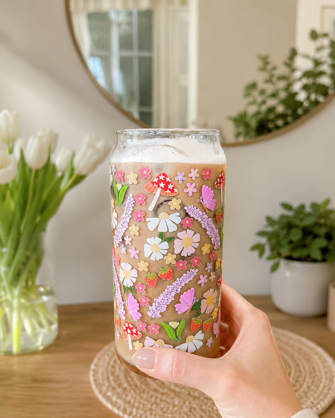 A hand holds a clear glass can cooler decorated with a colorful wildflower and mushroom pattern, filled with an iced coffee in a bright indoor setting with a mirror, tulips, and potted plants.