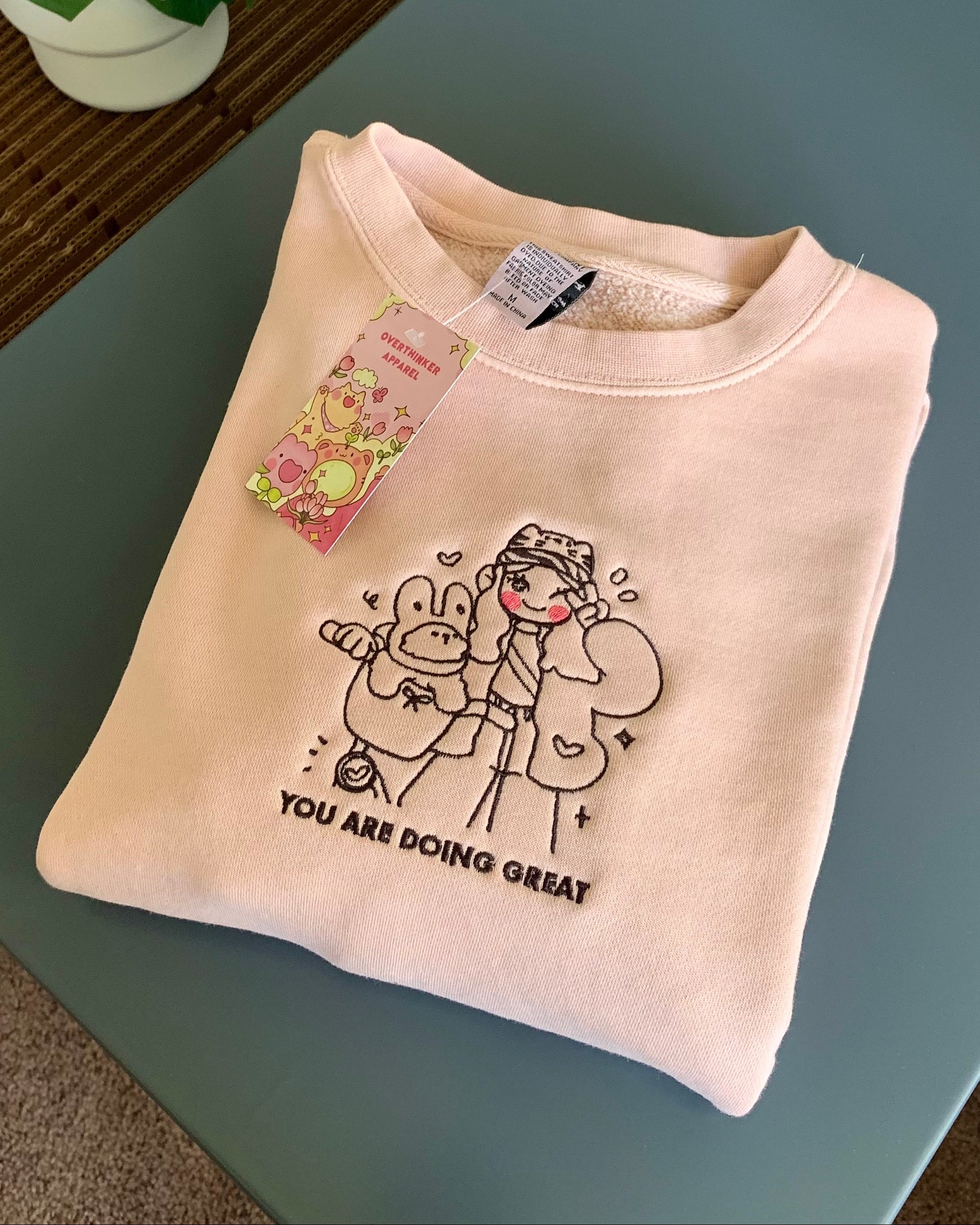 Folded dusty pink embroidered sweatshirt with “You Are Doing Great” message styled on a table with greenery