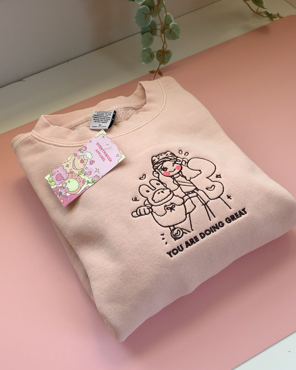 Dusty pink embroidered crewneck sweatshirt with a gentle illustrated design and “You Are Doing Great” text on a soft pink background