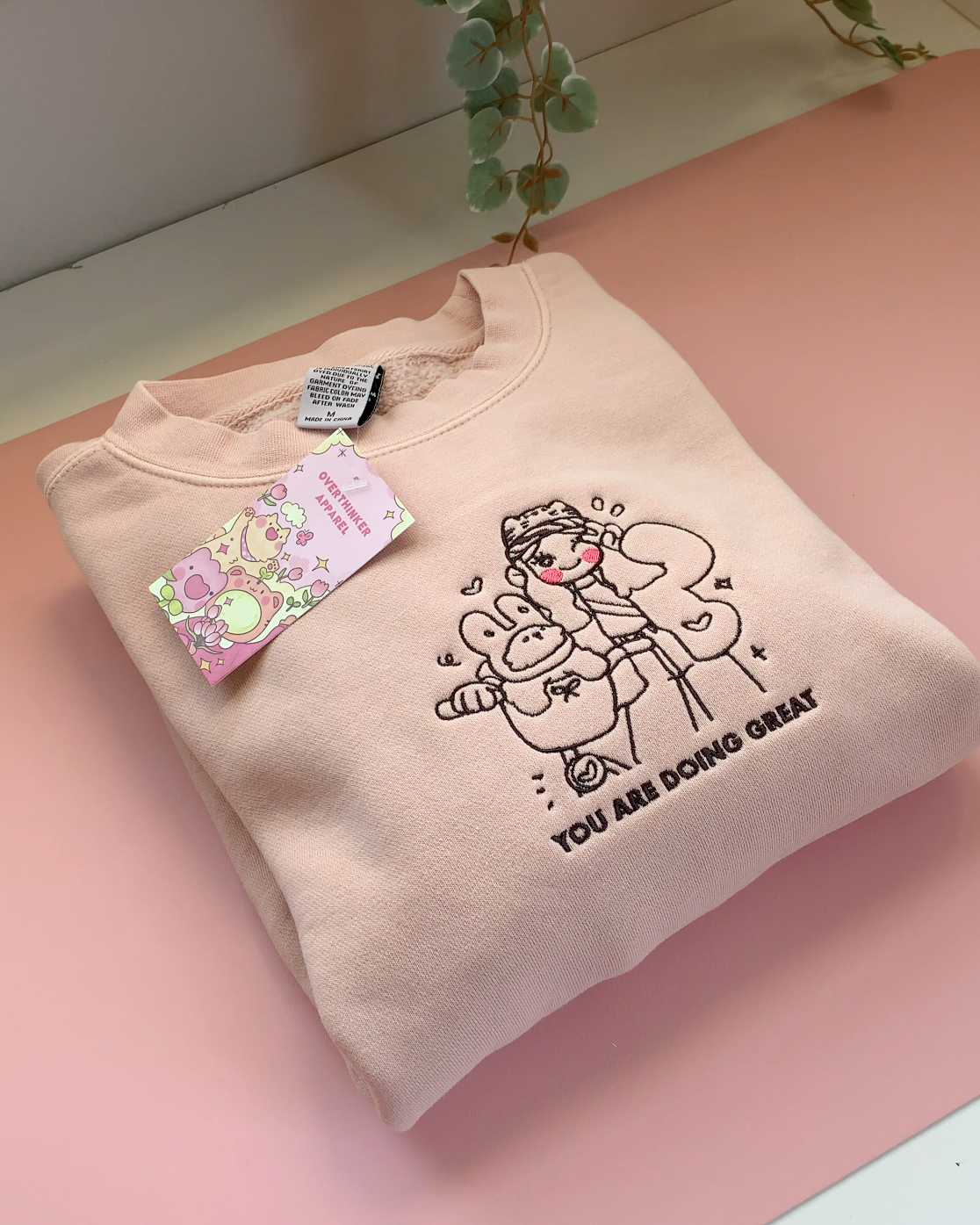 Dusty pink embroidered crewneck sweatshirt with a gentle illustrated design and “You Are Doing Great” text on a soft pink background