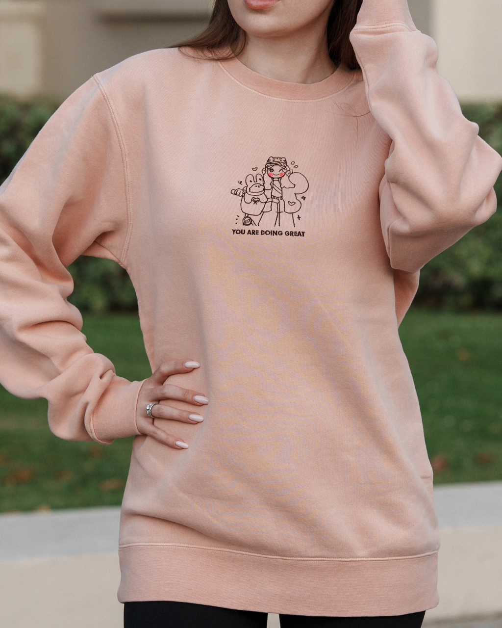 Model wearing a dusty pink embroidered crewneck sweatshirt with a small illustration and the words “You Are Doing Great,” designed as a soft, comforting affirmation sweatshirt for emotional support and everyday wear.
