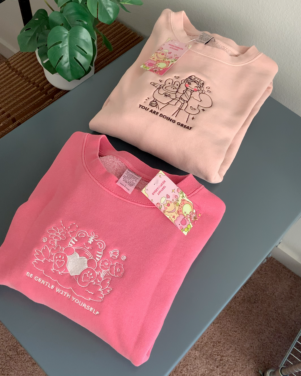 Dusty pink embroidered crewneck sweatshirt with a comforting illustrated duo and the words “You Are Doing Great,” folded flat on a table