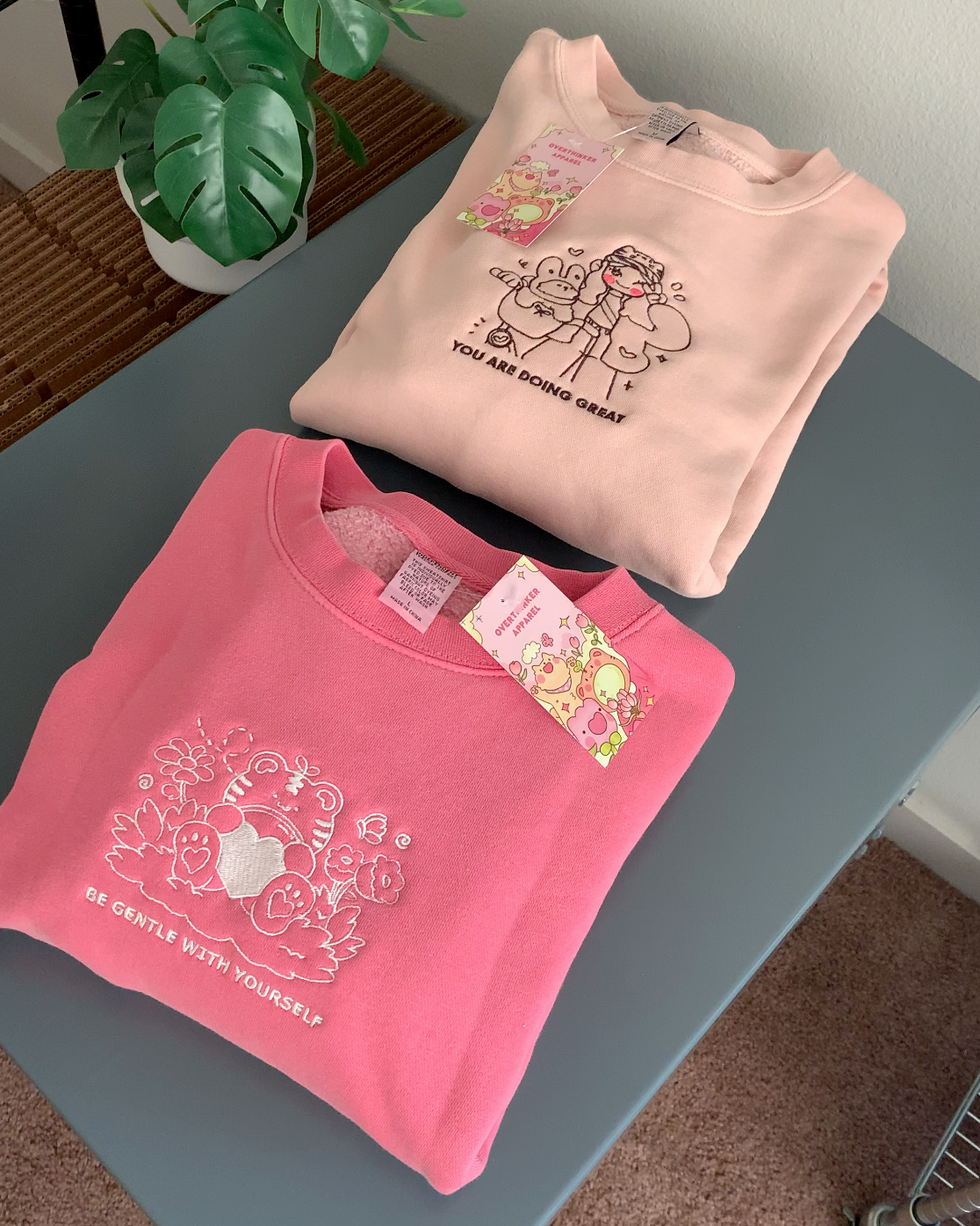 Dusty pink embroidered crewneck sweatshirt with a comforting illustrated duo and the words “You Are Doing Great,” folded flat on a table