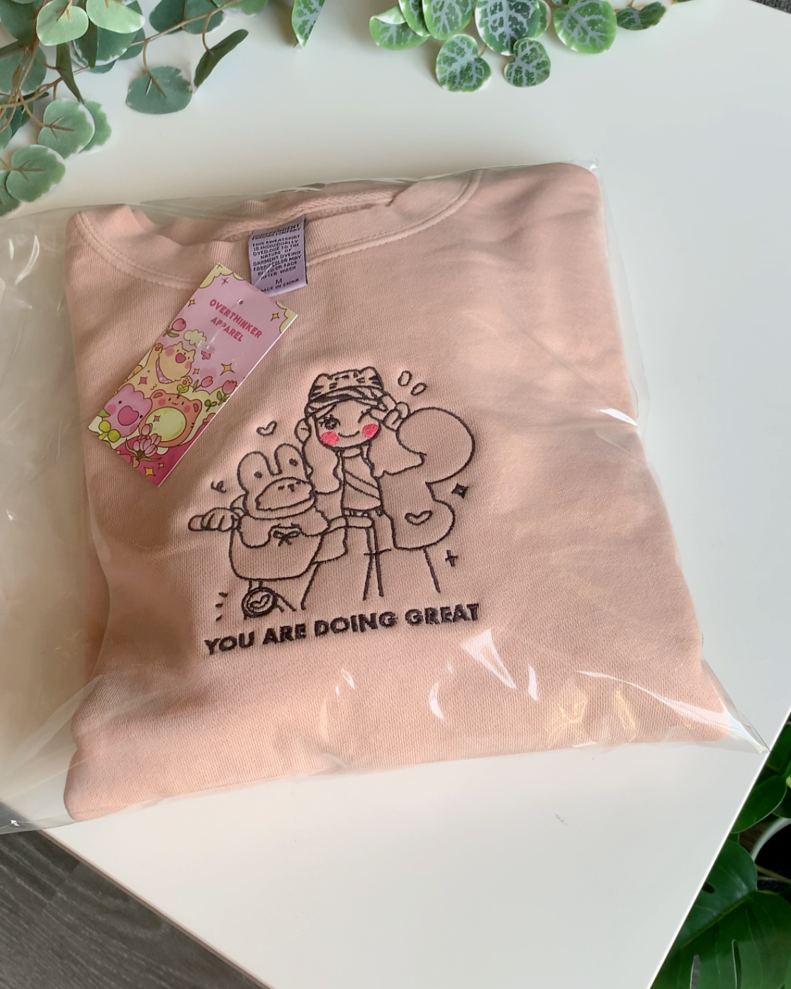 Top-down view of a dusty pink embroidered “You Are Doing Great” sweatshirt folded neatly in protective packaging