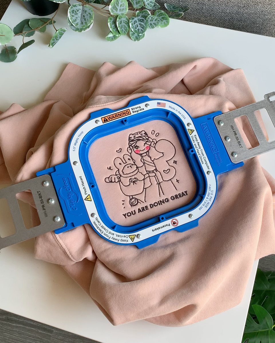 Close-up of hand-embroidered “You Are Doing Great” illustration stitched on a dusty pink sweatshirt in an embroidery hoop