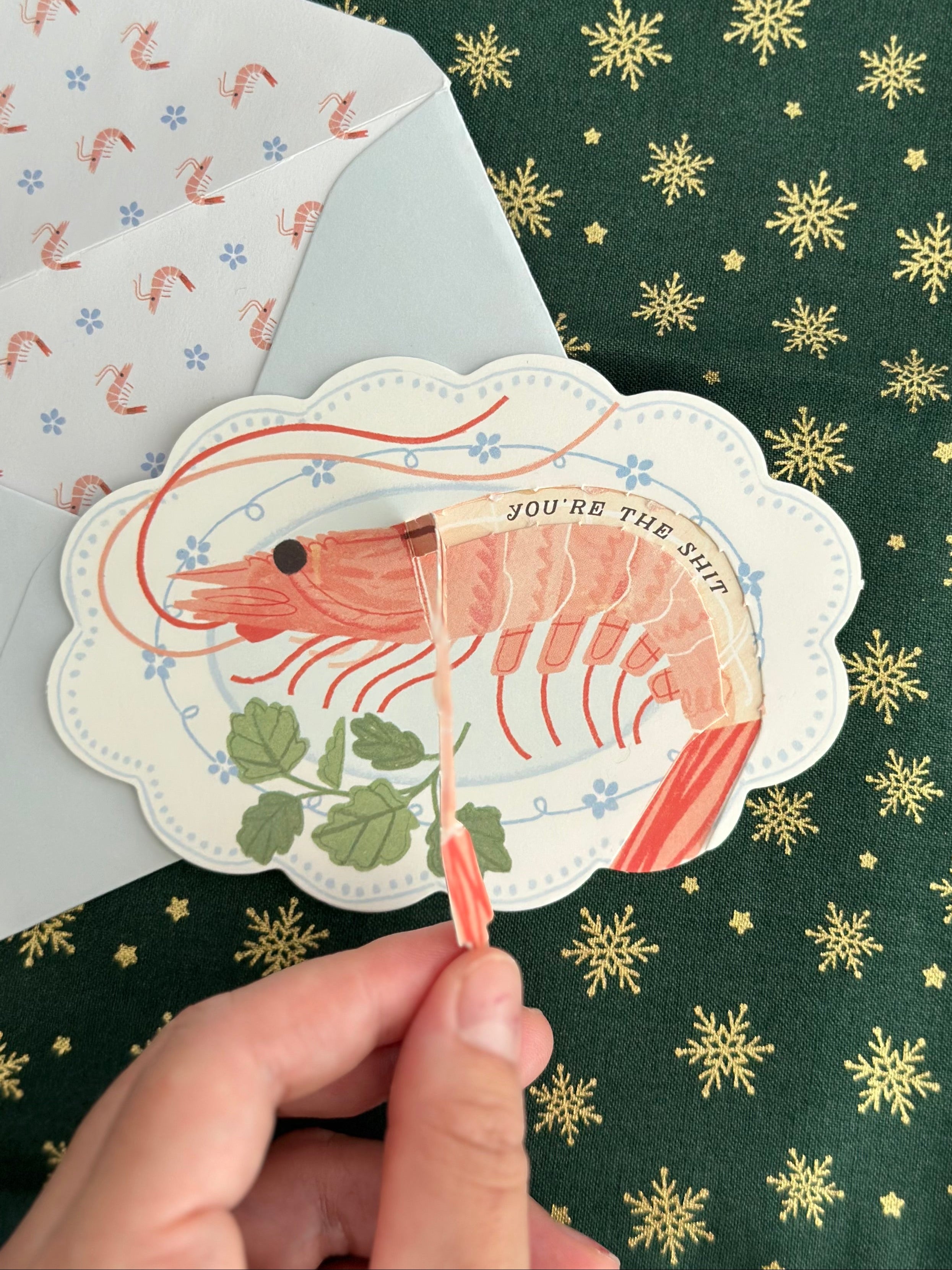 Hand pulling the red shrimp tail tab on a die-cut card to reveal hidden message; envelope with shrimp-patterned liner in background.