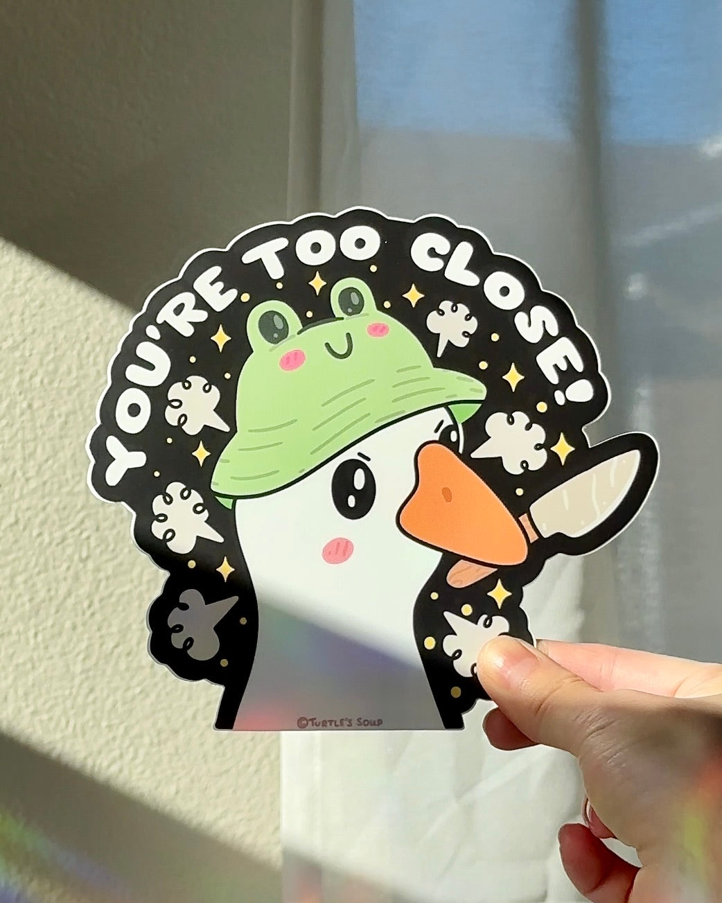 Hand holding a funny vinyl sticker with a goose wearing a frog hat and the text “You’re Too Close,” humorous boundary and personal space car decal.
