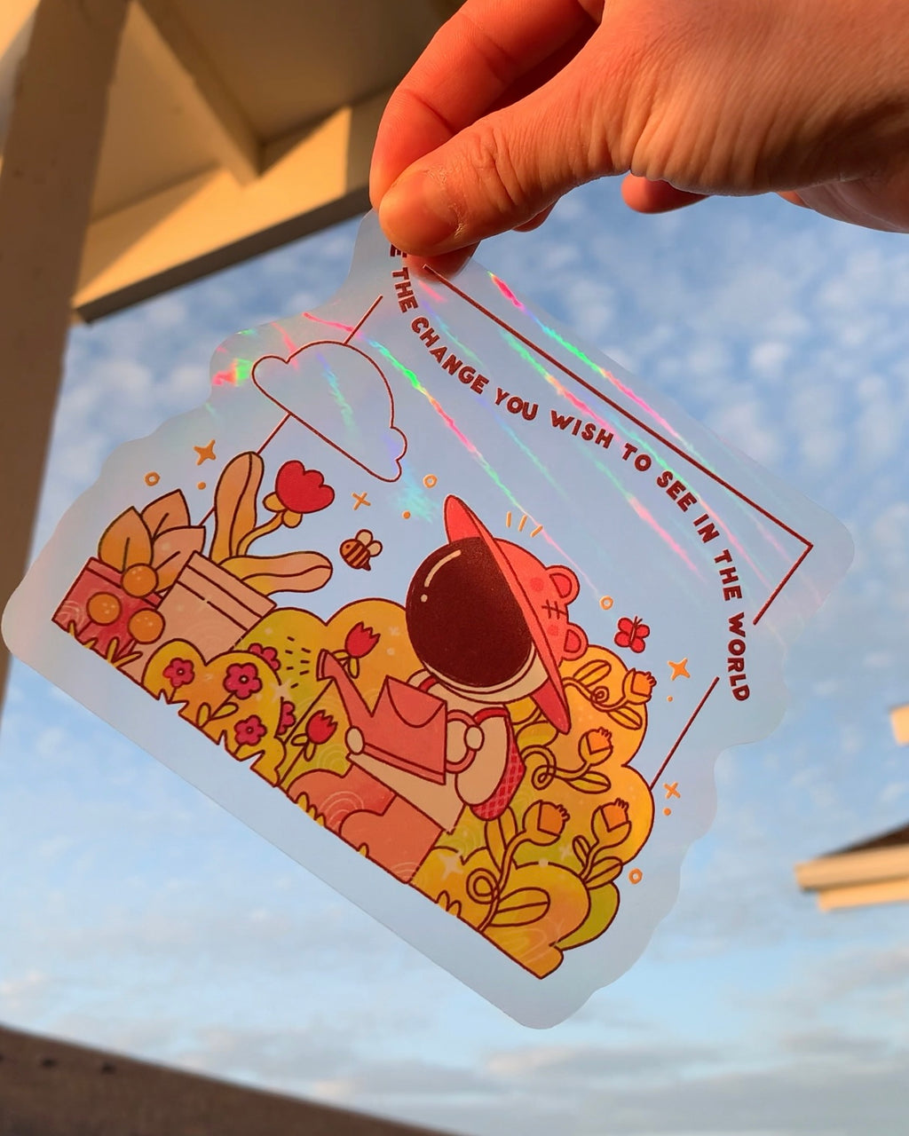 Hand holding a rainbow window suncatcher static cling sticker in sunlight, featuring a garden illustration and inspirational message that creates soft rainbow light.